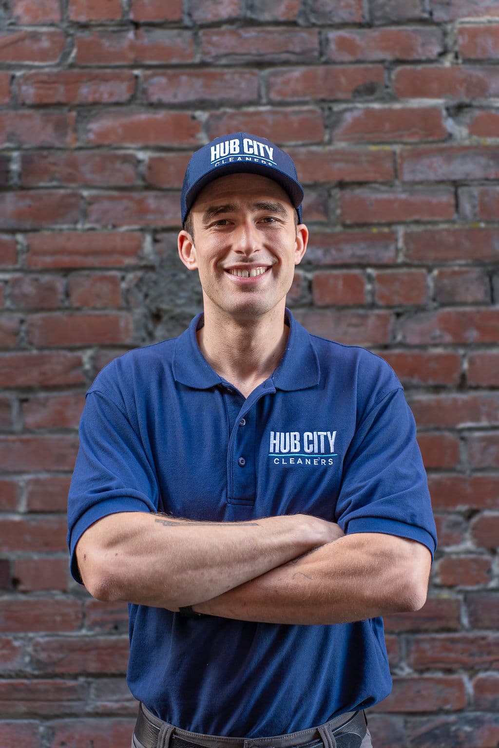 Caleb smiling in a Hub City Cleaners polo and cap in front of a brick wall